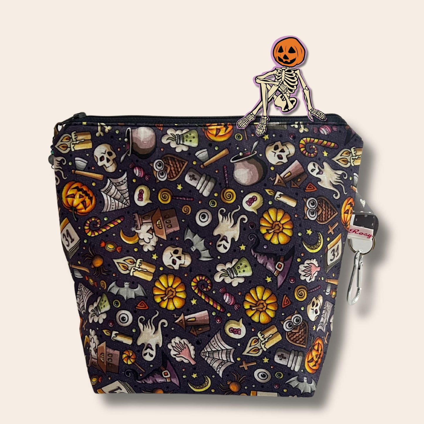 A dark grey bag covered in cartoon Halloween illustrations. A sticker of a skeleton with a pumpkin head sits on top of the bag.