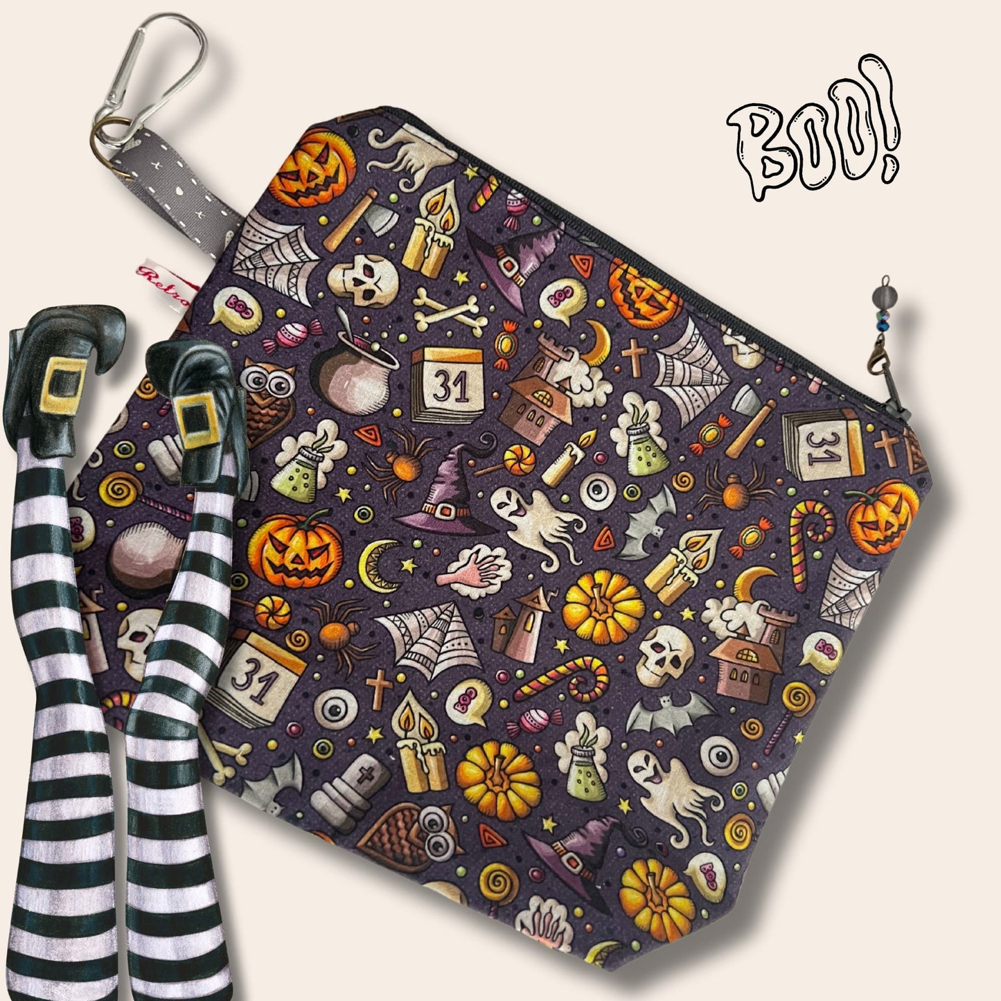A dark grey bag covered in cartoon Halloween illustrations. Bubble text reads "Boo!". A pair of upside-down legs with pointy buckled shoes and striped tights.